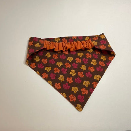 A scrunchie dog bandana with a chocolate background and an all-over pattern of small orange, yellow and red maple leaves.