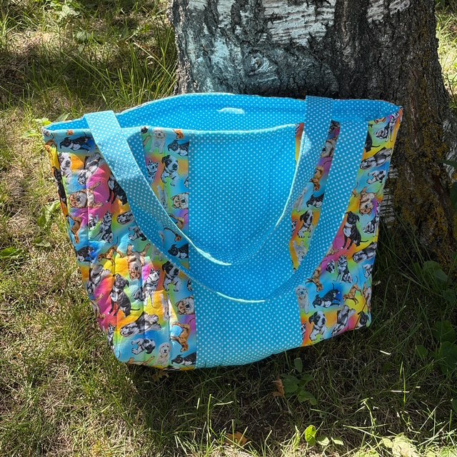 A large tote bag with a bright blue polka-dot front panel and colorful cartoon character side panels, leaning against a tree trunk on grass outdoors.