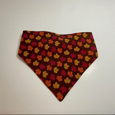 A scrunchie dog bandana with a chocolate background and an all-over pattern of small orange, yellow and red maple leaves.