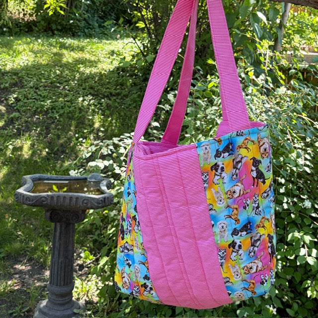 A handmade fabric tote bag with bright pink and multi dog themed fabric panels and handles hanging outside in a garden.