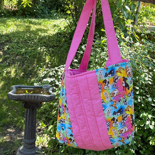 A handmade fabric tote bag with bright pink and multi dog themed fabric panels and handles hanging outside in a garden.