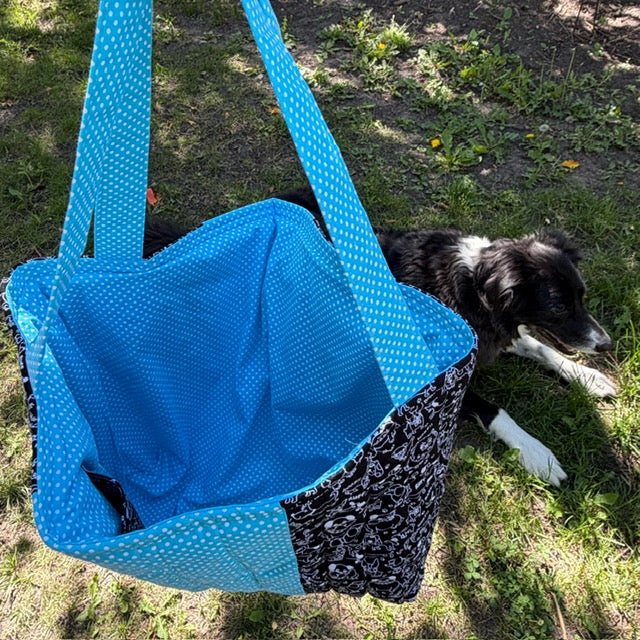 A large fabric tote bag hanging  - the bag has bright blue polka-dot shoulder straps and front pocket, with black and white dog themed panels -  shown outdoors on grass next to Buddy the Border Collie.