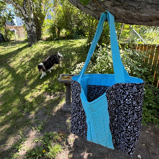 A large fabric tote bag hanging  - the bag has bright blue polka-dot shoulder straps and front pocket, with black and white dog themed panels.