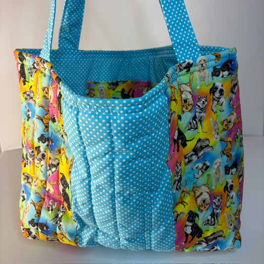 A handmade quilted tote bag featuring a central light blue and white polka dot panel, flanked by two colorful side panels printed with various dog illustrations. The bag has two shoulder straps made of the blue polka dot fabric.